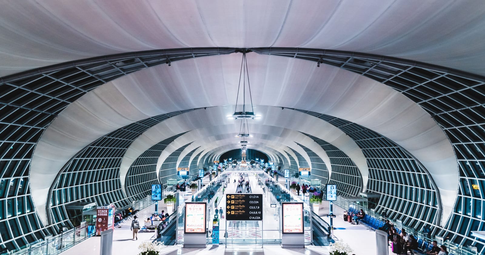 The arched glass concourse of Bangkok's Suvarnabhumi Airport at night, with travellers walking to their gates